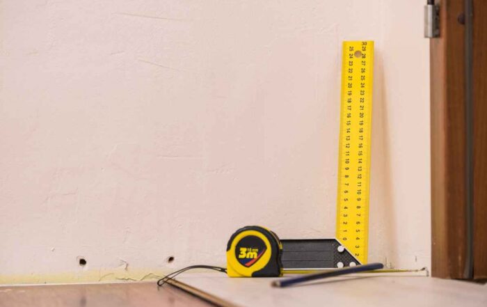 A close-up photograph of a yellow L-square ruler and a tape measure placed against a light-colored interior wall to measure a gap between the wall and the flooring.
