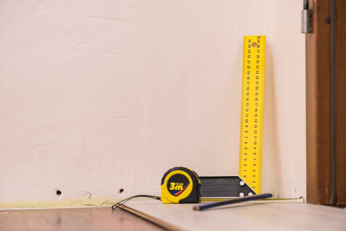 A close-up photograph of a yellow L-square ruler and a tape measure placed against a light-colored interior wall to measure a gap between the wall and the flooring.